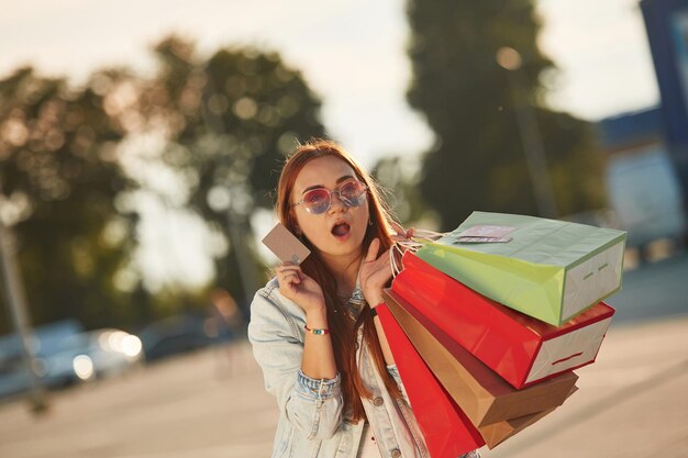 Photo with credit card beautiful woman in casual clothes is holding shopping bags outdoors