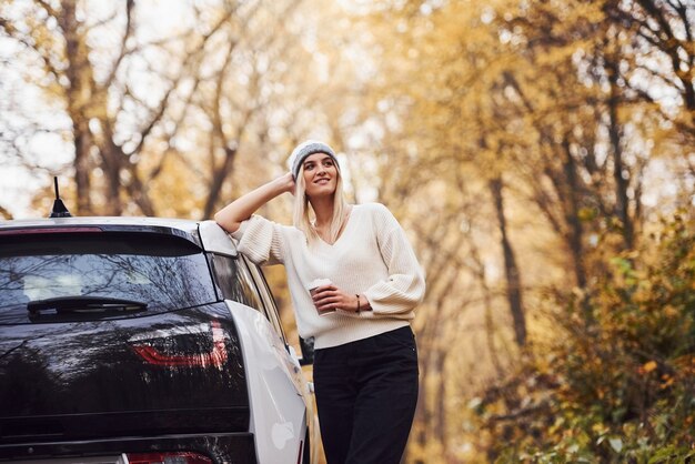 With cup of drink in hands. Girl have autumn trip by car. Modern brand new automobile in the forest.