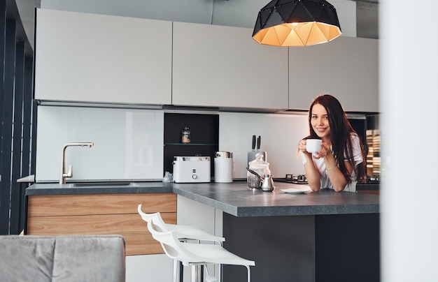 With cup of fresh coffee Young beautiful brunette in casual clothes indoors in kitchen at daytime