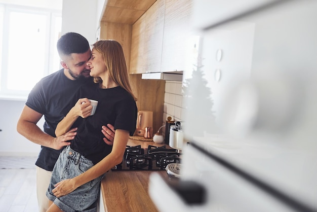 With cup of fresh drink. Morning for young married couple that standing indoors in the kitchen.