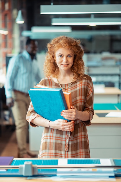 With curly hair. Beautiful red-haired woman with curly hair holding books standing in publishing office