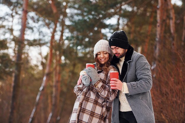 With drink Cheerful couple have a walk in the winter forest at daytime