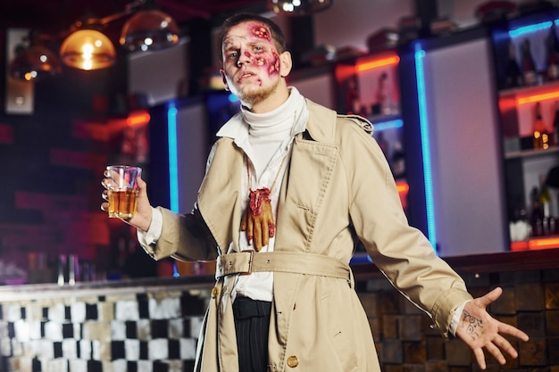 With drink in hand. Portrait of man that is on the thematic halloween party in zombie makeup and costume.