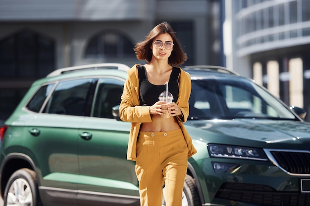 With drink in hands Young fashionable woman in burgundy colored coat at daytime with her car
