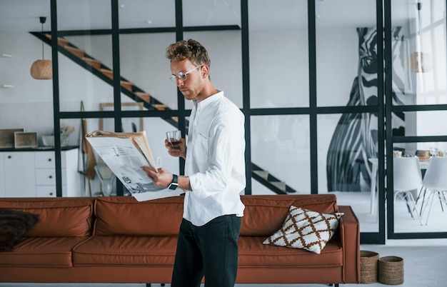 With drink and newspaper Caucasian young guy in elegant white shirt indoors at home