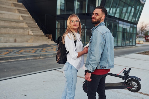 With electric schooter Young stylish man with woman in casual clothes outdoors near business building together Conception of friendship or relationships