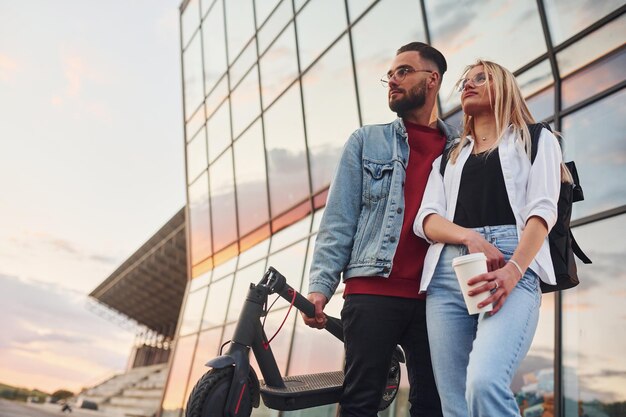 With electric schooter Young stylish man with woman in casual clothes outdoors near business building together Conception of friendship or relationships