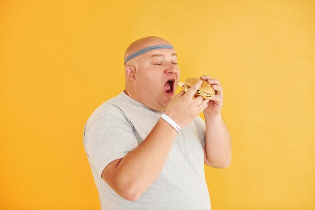 Photo with fast food funny overweight man in sportive head tie is against yellow background