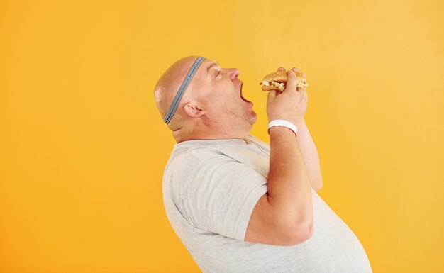 With fast food Funny overweight man in sportive head tie is against yellow background