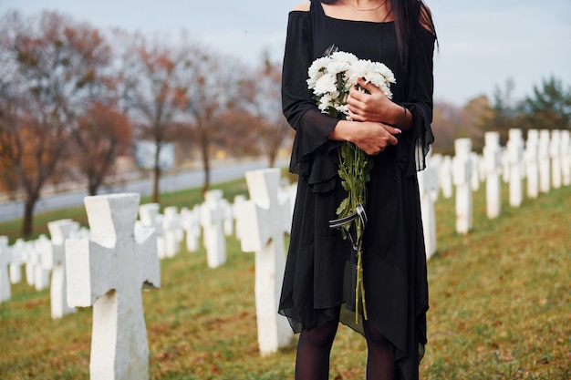 With flowers in hands young woman in black clothes visiting\
cemetery with many white crosses conception of funeral and\
death