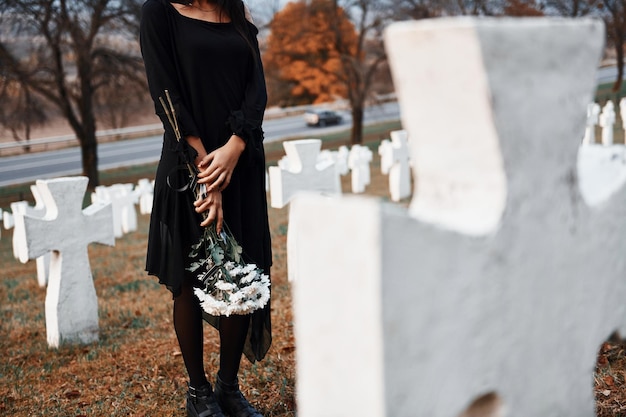 Photo with flowers in hands young woman in black clothes visiting cemetery with many white crosses conception of funeral and death