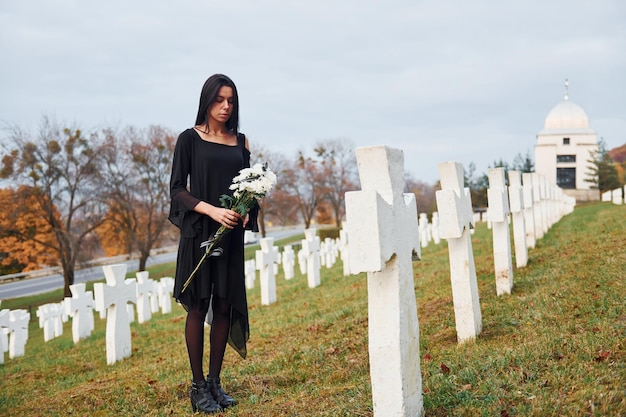 With flowers in hands young woman in black clothes visiting\
cemetery with many white crosses conception of funeral and\
death