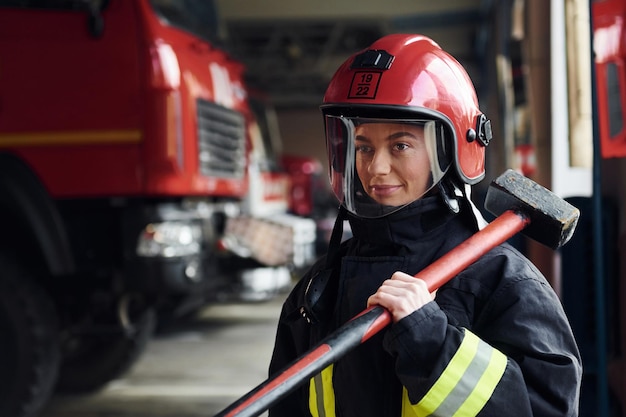 With hammer in hands female firefighter in protective uniform\
standing near truck