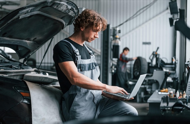 With laptop Adult man in grey colored uniform works in the automobile salon