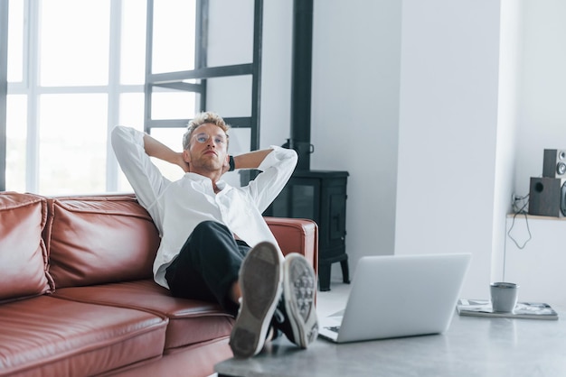 With laptop Caucasian young guy in elegant white shirt indoors at home