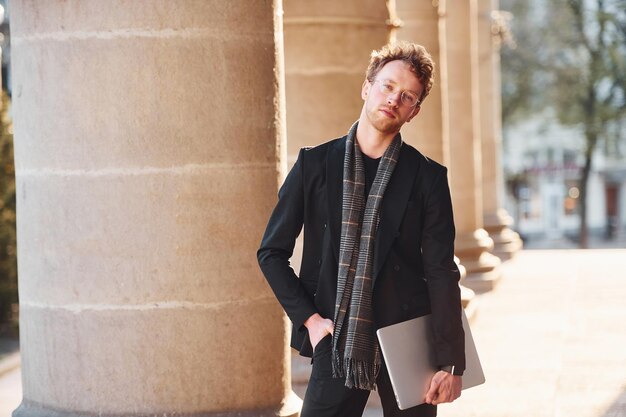 Con il laptop in mano elegante giovane uomo con gli occhiali e in abiti formali di classe all'aperto in città