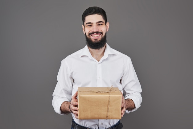 With love for you. Good looking young man in blue jeans shirt holding a gift box and looking at camera while standing on grey