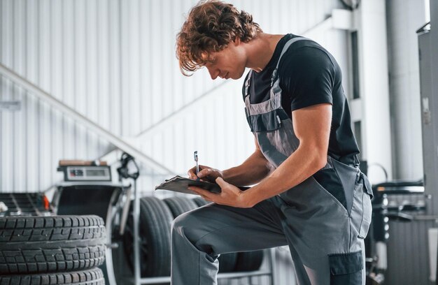 With notepad in hands Adult man in grey colored uniform works in the automobile salon