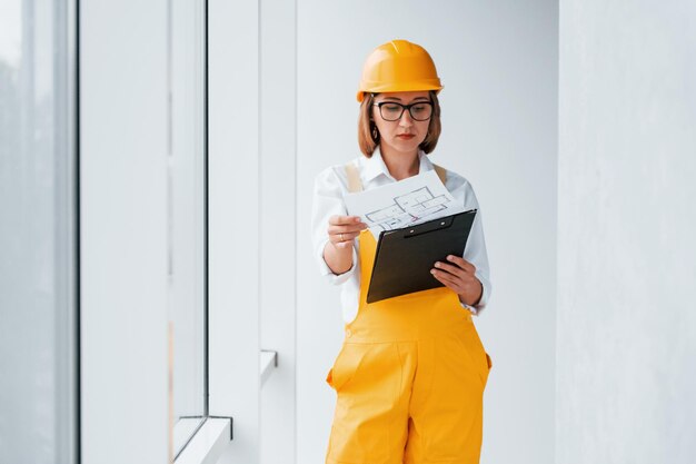 With notepad in hands Female worker or engineer in yellow uniform and hard hat standing indoors