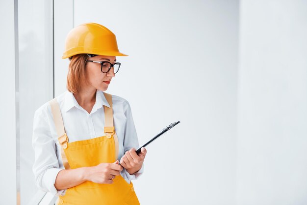 With notepad in hands Female worker or engineer in yellow uniform and hard hat standing indoors