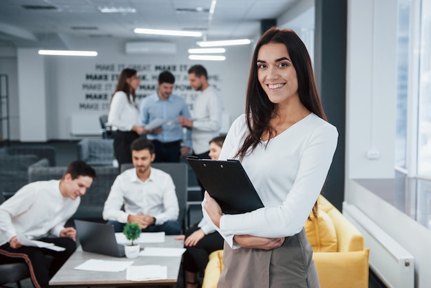 With notepad in hands. Portrait of young girl stands in the office with employees at background