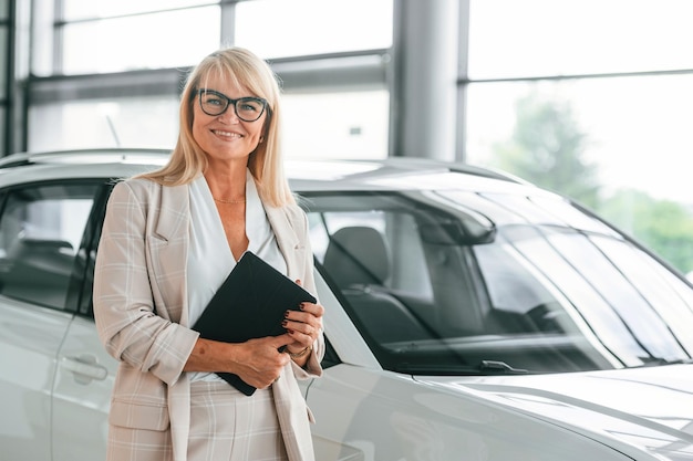 With notepad in hands woman in white formal clothes is in the\
car dealership