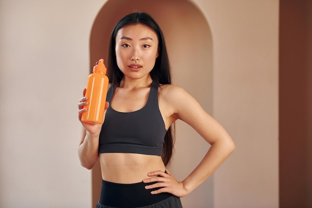 With orange colored bottle Young serious asian woman standing indoors