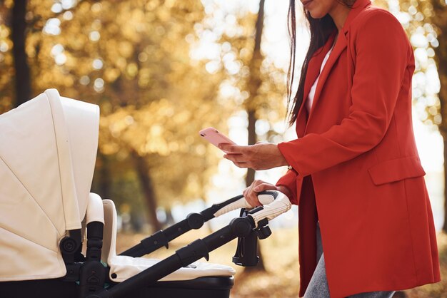 With phone in hand. Mother in red coat have a walk with her kid in the pram in the park at autumn time.