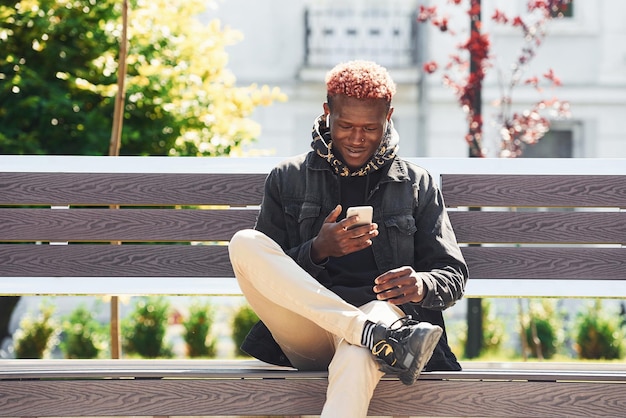 With phone in hand Young african american man in black jacket outdoors in the city sitting on bench