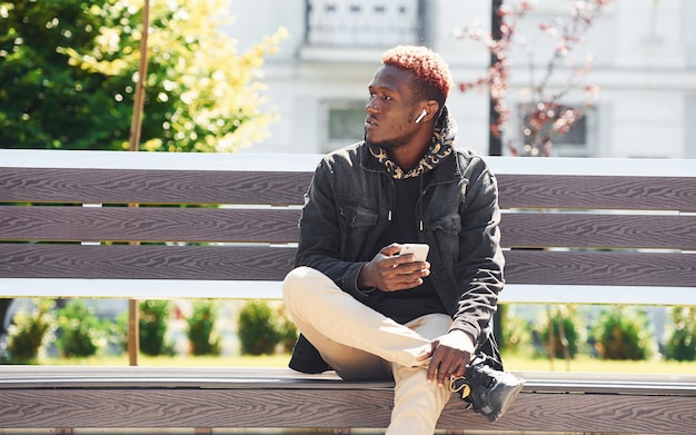 With phone in hand Young african american man in black jacket outdoors in the city sitting on bench