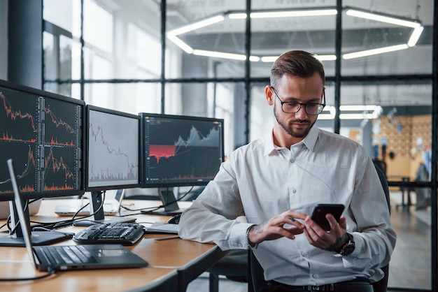 With phone in hands. Male stockbroker in formal clothes works in the office with financial market.