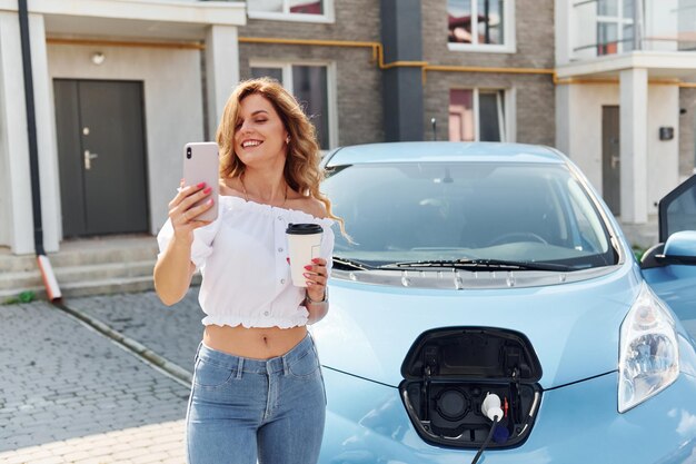 Photo with phone young woman in casual clothes with her electromobile outdoors at daytime