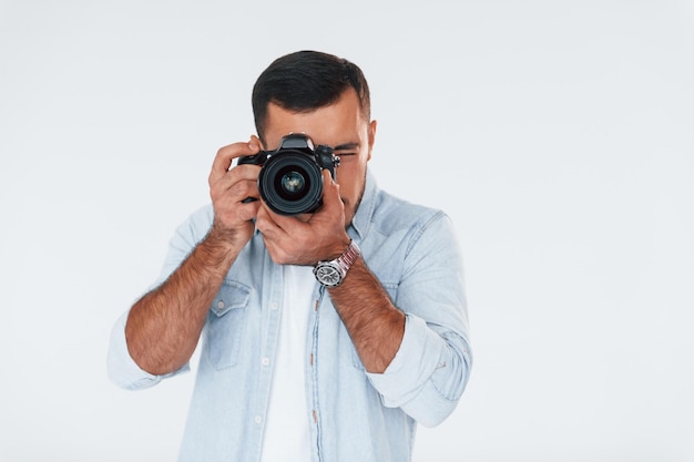 Con fotocamera professionale giovane bell'uomo in piedi al chiuso su sfondo bianco