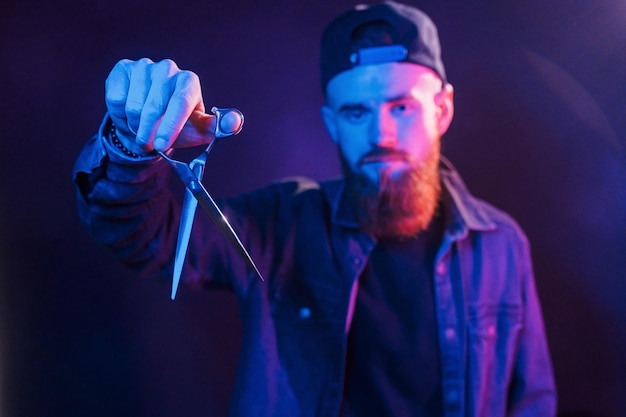 With scissors Young bearded barber in cap standing in the studio with neon lighting