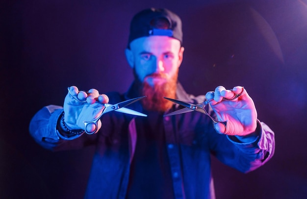 With scissors Young bearded barber in cap standing in the studio with neon lighting