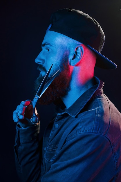 With scissors Young bearded barber in cap standing in the studio with neon lighting