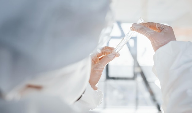 With test tube in hand Female doctor scientist in lab coat defensive eyewear and mask standing indoors