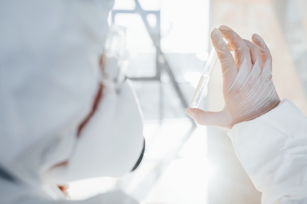 Con la provetta in mano. scienziato medico femminile in camice da laboratorio, occhiali difensivi e maschera in piedi al chiuso