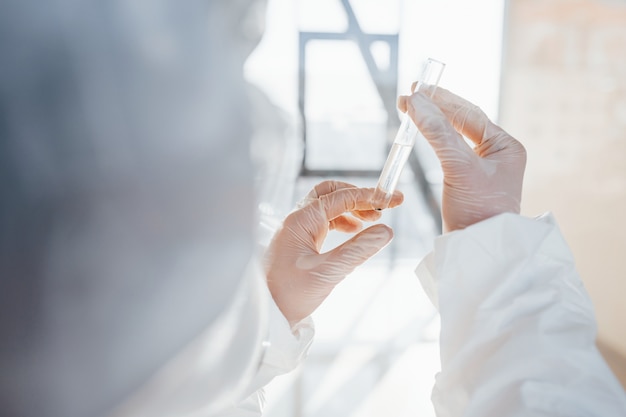with test tube in hand. female doctor scientist in lab coat, defensive eyewear and mask standing indoors