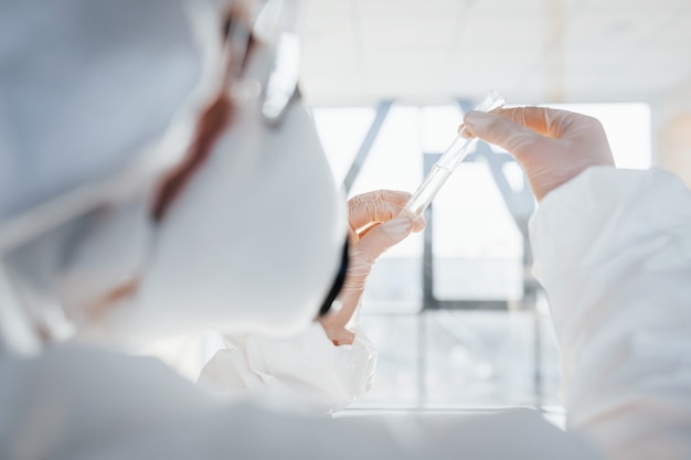 Con la provetta in mano. scienziato medico femminile in camice da laboratorio, occhiali difensivi e maschera in piedi al chiuso