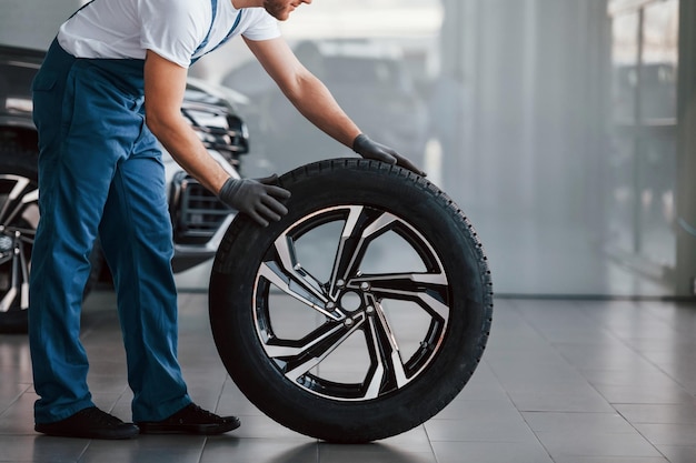 With tire Young man in white shirt and blue uniform repairs automobile