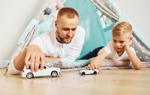 With toy cars Father and his young young spending time together indoors near tent