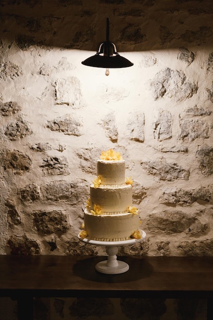 Foto witte cake met drie niveaus op een standaard tegen een achtergrond van een stenen muur onder een lamp