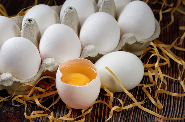 Witte eieren in een pak op papier stro en houten tafel. Gebroken ei met dooier aan de voorkant. Low key.