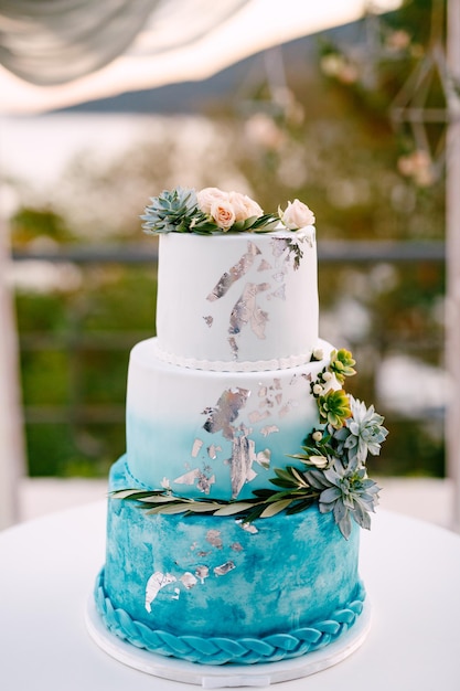 Witte en blauwe bruidstaart met bloemen en vetplanten staat op een cakestand op een witte tafel