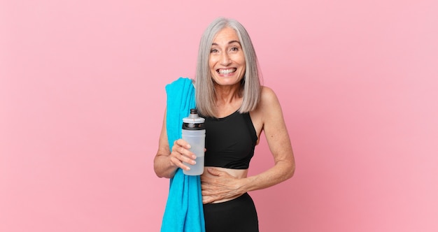 Witte haarvrouw van middelbare leeftijd die hardop lacht om een hilarische grap met een handdoek en een waterfles. fitnessconcept