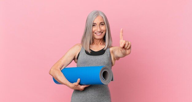 Witte haarvrouw van middelbare leeftijd die lacht en er vriendelijk uitziet, nummer één toont en een yogamat-fitnessconcept vasthoudt