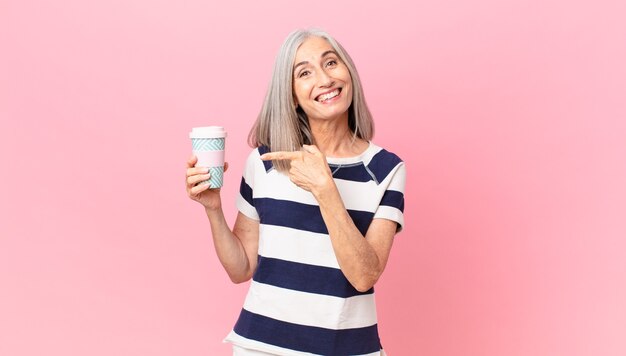 Witte haarvrouw van middelbare leeftijd die opgewonden en verrast kijkt en naar de zijkant wijst en een koffiebeker vasthoudt