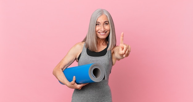 Witte haarvrouw van middelbare leeftijd die trots en zelfverzekerd glimlacht en nummer één maakt en een yogamat vasthoudt. fitness concept