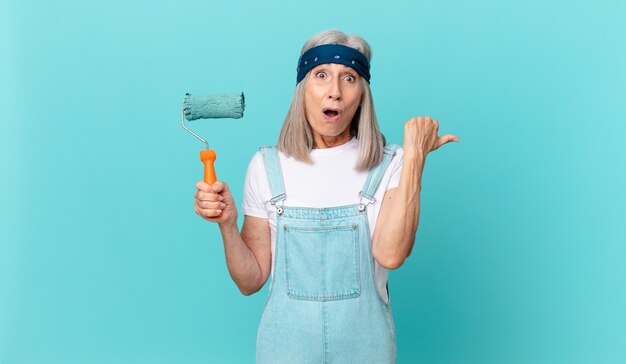 Witte haarvrouw van middelbare leeftijd die verbaasd kijkt in ongeloof met een roller die een muur schildert